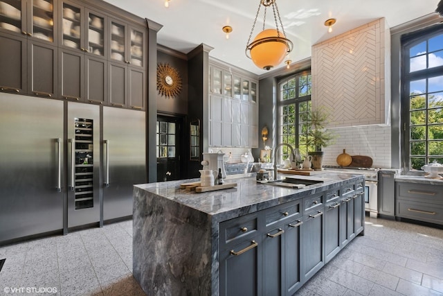 kitchen featuring hanging light fixtures, glass insert cabinets, a sink, an island with sink, and dark stone counters