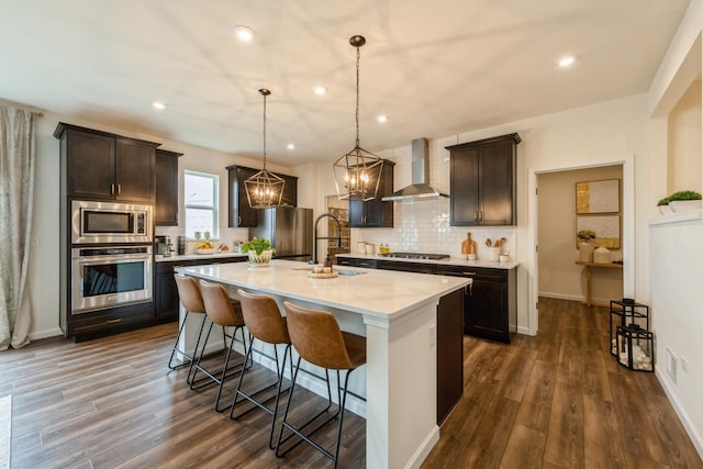 kitchen with appliances with stainless steel finishes, wall chimney exhaust hood, an island with sink, pendant lighting, and a kitchen bar