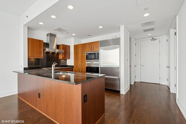 kitchen featuring dark stone counters, wall chimney range hood, built in appliances, dark hardwood / wood-style floors, and kitchen peninsula