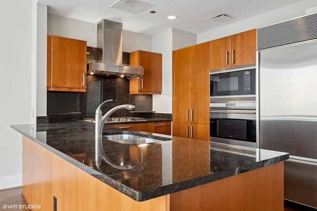 kitchen featuring tasteful backsplash, wall chimney range hood, built in appliances, dark stone countertops, and an island with sink