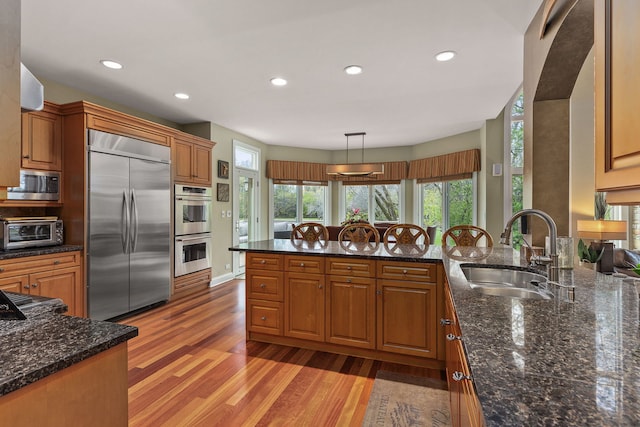 kitchen featuring pendant lighting, sink, built in appliances, dark stone counters, and light wood-type flooring