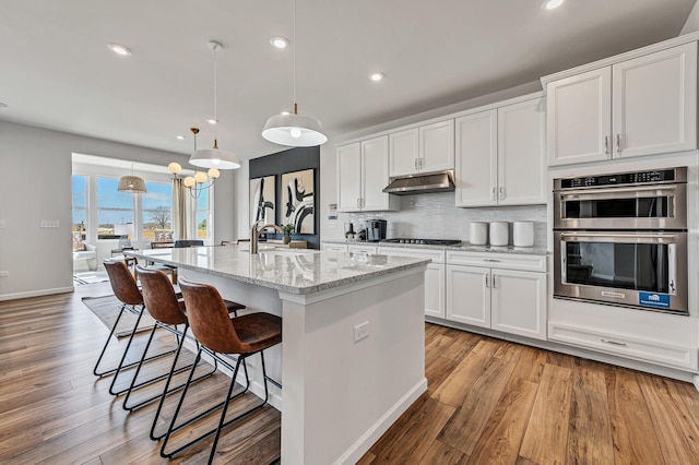 kitchen featuring light stone countertops, stainless steel double oven, decorative light fixtures, a kitchen island with sink, and white cabinets