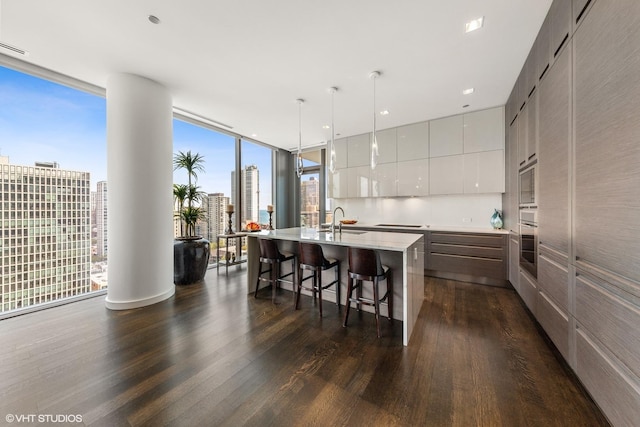 kitchen with a center island with sink, sink, hanging light fixtures, white cabinetry, and a wall of windows