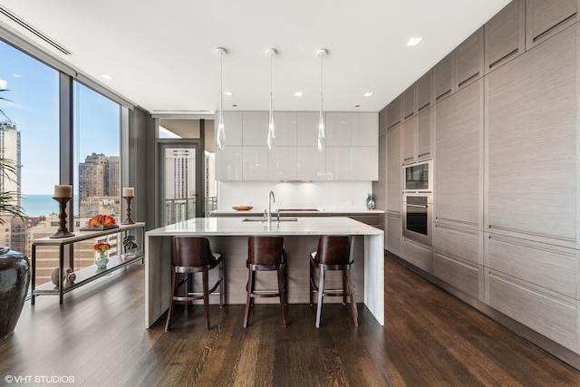 kitchen featuring a wall of windows, an island with sink, pendant lighting, oven, and white cabinets