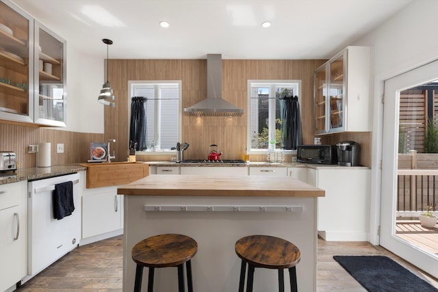 kitchen with a kitchen bar, white dishwasher, wall chimney range hood, pendant lighting, and white cabinets