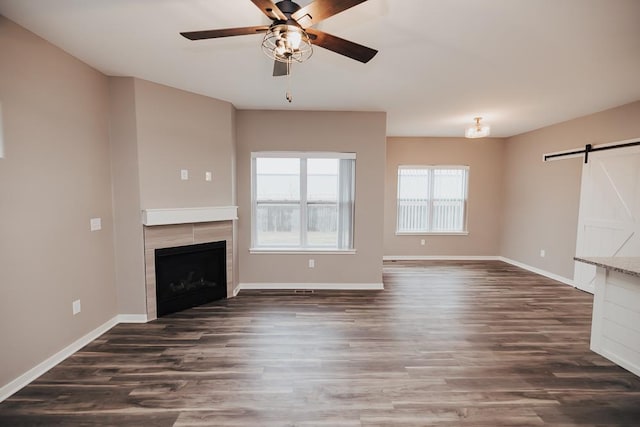 unfurnished living room with ceiling fan, a barn door, dark hardwood / wood-style flooring, and a tile fireplace