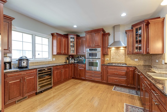 kitchen featuring wall chimney exhaust hood, stainless steel appliances, wine cooler, light hardwood / wood-style flooring, and dark stone countertops