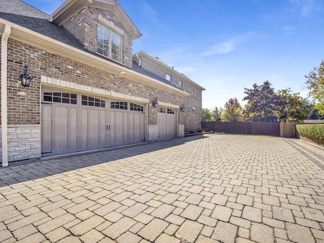 view of property exterior with a garage, a shingled roof, stone siding, fence, and brick siding