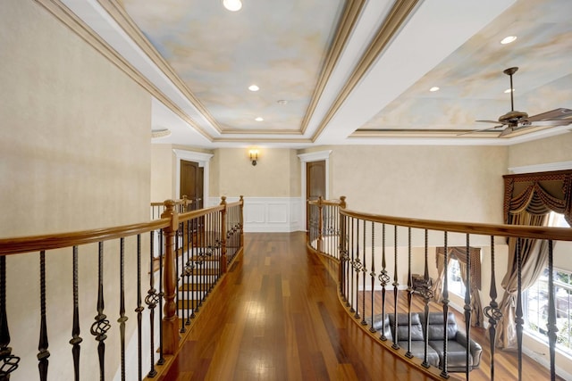 corridor with a wainscoted wall, a raised ceiling, wood-type flooring, a decorative wall, and an upstairs landing