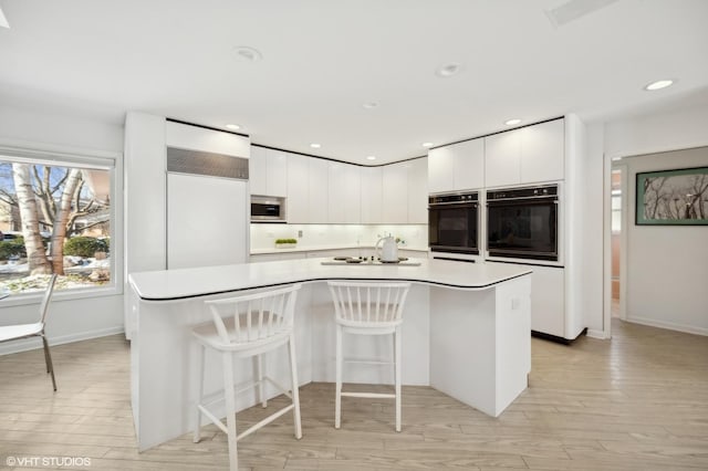 kitchen with a center island with sink, a breakfast bar area, double oven, white cabinets, and light hardwood / wood-style flooring