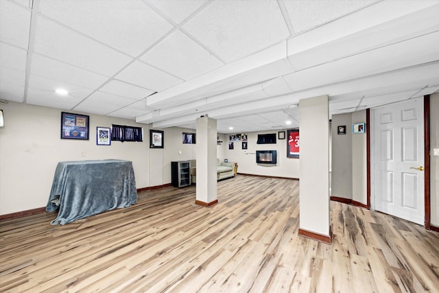 finished basement with a glass covered fireplace, a paneled ceiling, baseboards, and light wood finished floors