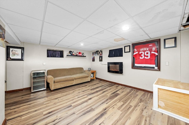 living area featuring light wood finished floors, baseboards, wine cooler, a glass covered fireplace, and a paneled ceiling