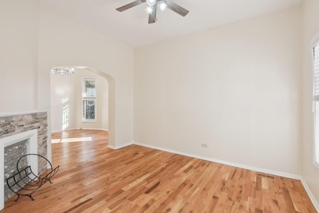 unfurnished living room with a stone fireplace, ceiling fan with notable chandelier, and light wood-type flooring