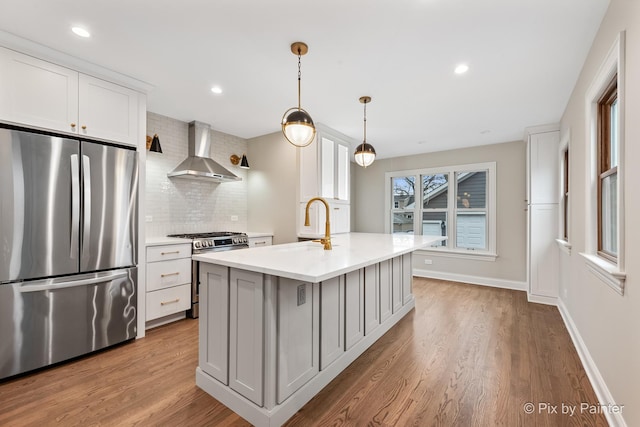 kitchen with white cabinets, hanging light fixtures, a kitchen island with sink, stainless steel appliances, and wall chimney range hood