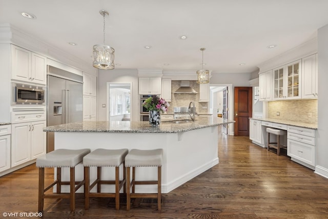 kitchen with white cabinetry, wall chimney range hood, built in appliances, an island with sink, and decorative light fixtures