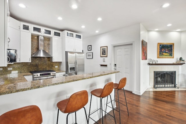 kitchen featuring wall chimney range hood, kitchen peninsula, decorative backsplash, appliances with stainless steel finishes, and white cabinetry