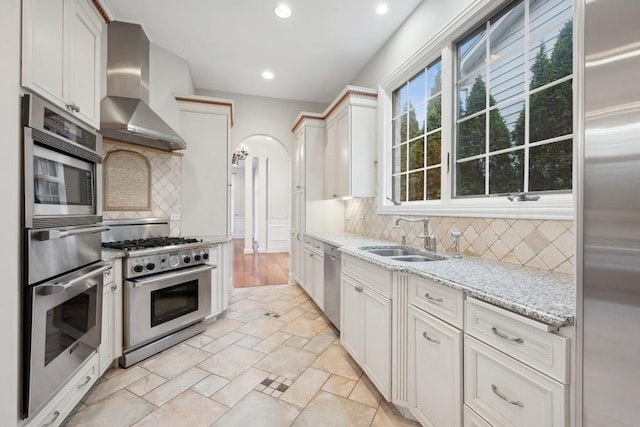 kitchen featuring light stone countertops, sink, stainless steel appliances, wall chimney range hood, and white cabinets