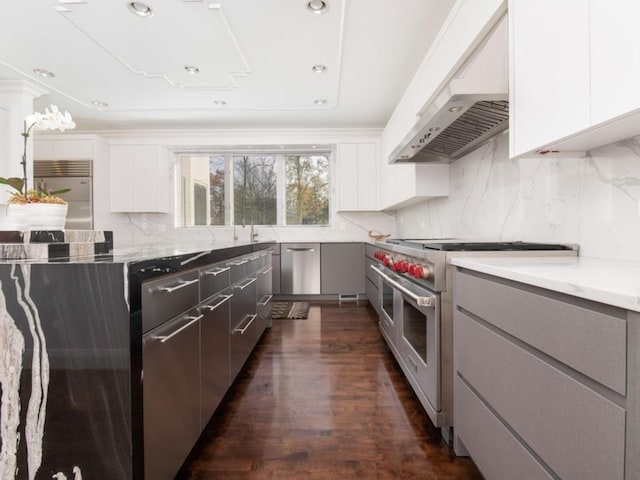 kitchen featuring high end appliances, dark wood-type flooring, white cabinets, wall chimney range hood, and hanging light fixtures