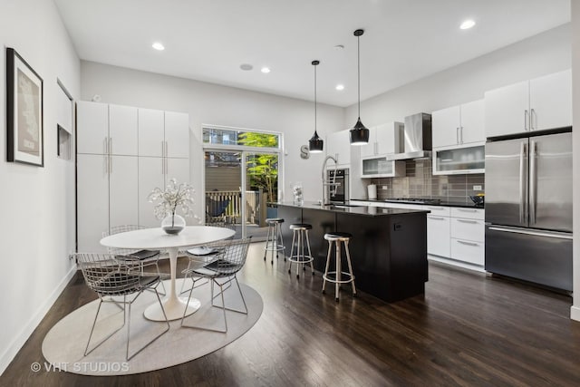 kitchen with white cabinetry, wall chimney exhaust hood, an island with sink, backsplash, and appliances with stainless steel finishes