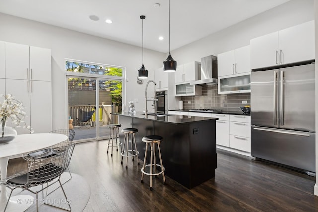 kitchen with hanging light fixtures, wall chimney range hood, a kitchen island with sink, backsplash, and appliances with stainless steel finishes