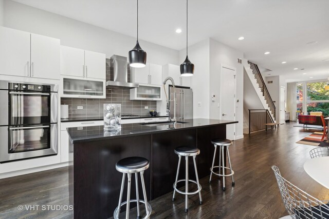 kitchen featuring a breakfast bar area, stainless steel double oven, a kitchen island with sink, and wall chimney exhaust hood