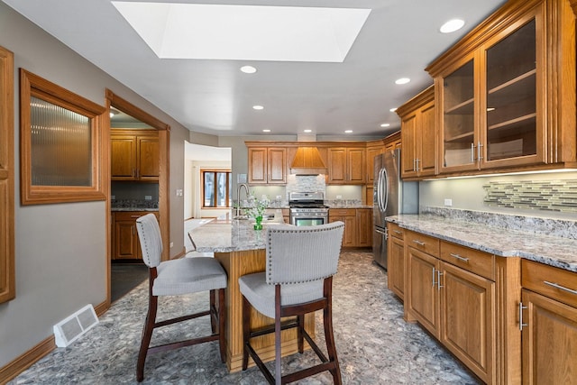 kitchen with visible vents, brown cabinetry, appliances with stainless steel finishes, a kitchen island with sink, and a kitchen bar