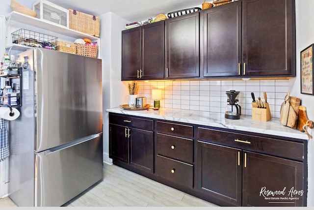 kitchen with backsplash, stainless steel refrigerator, dark brown cabinetry, and light stone counters