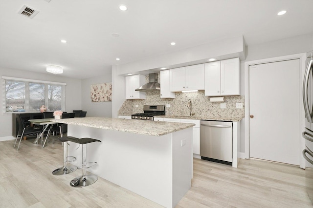 kitchen with white cabinetry, stainless steel appliances, wall chimney range hood, light stone counters, and a kitchen island