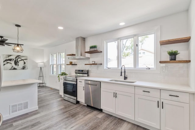 kitchen featuring appliances with stainless steel finishes, wall chimney range hood, sink, decorative light fixtures, and white cabinets