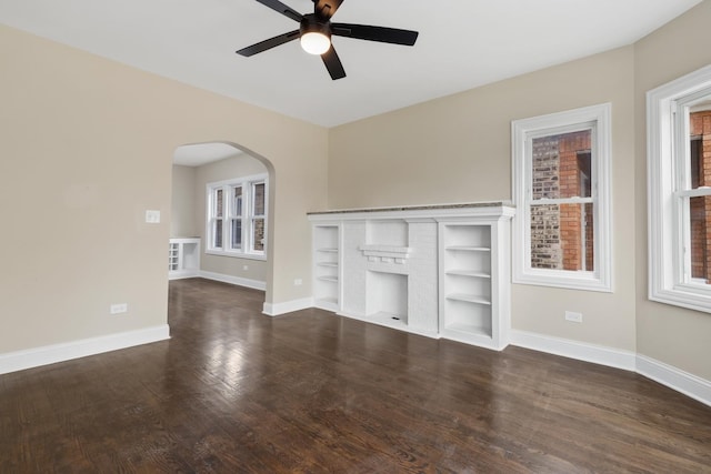 unfurnished living room featuring built in shelves, dark hardwood / wood-style floors, a brick fireplace, and ceiling fan