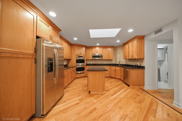 kitchen featuring a kitchen island, appliances with stainless steel finishes, a skylight, dark stone counters, and light hardwood / wood-style flooring