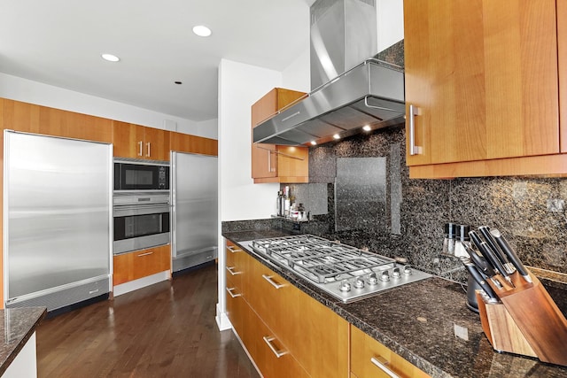 kitchen with built in appliances, decorative backsplash, dark stone countertops, and wall chimney range hood