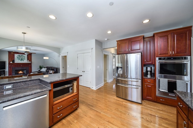 kitchen with appliances with stainless steel finishes, hanging light fixtures, light wood-type flooring, dark stone counters, and ceiling fan