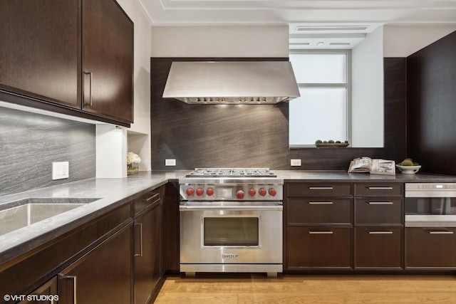 kitchen with dark brown cabinetry, wall chimney exhaust hood, premium range, oven, and stone countertops