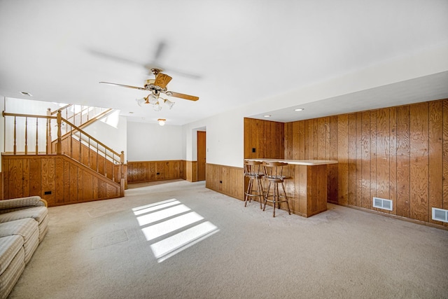 unfurnished living room with ceiling fan, light colored carpet, wooden walls, and bar area