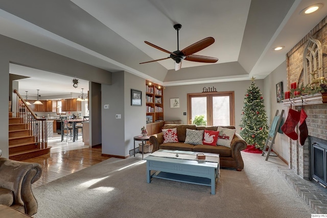 carpeted living room with a tray ceiling, ceiling fan with notable chandelier, and a brick fireplace