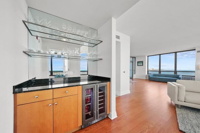 bar featuring a healthy amount of sunlight, wine cooler, dark stone countertops, light hardwood / wood-style floors, and a water view