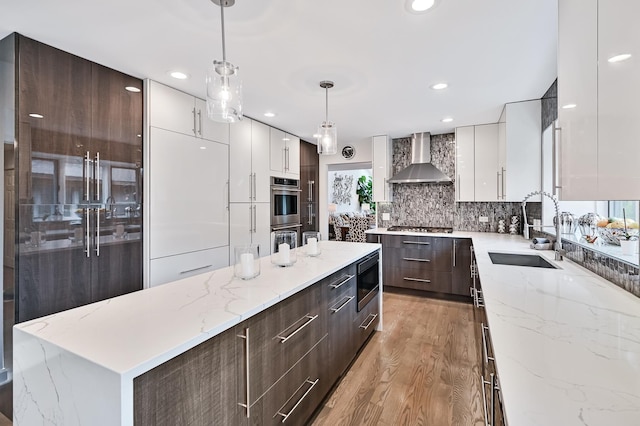 kitchen with a center island, wall chimney range hood, sink, hanging light fixtures, and light hardwood / wood-style flooring