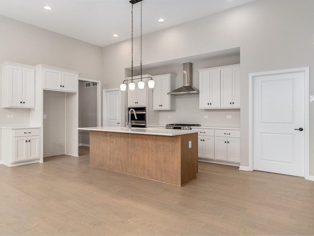 kitchen featuring a towering ceiling, wall chimney range hood, white cabinets, pendant lighting, and a center island with sink