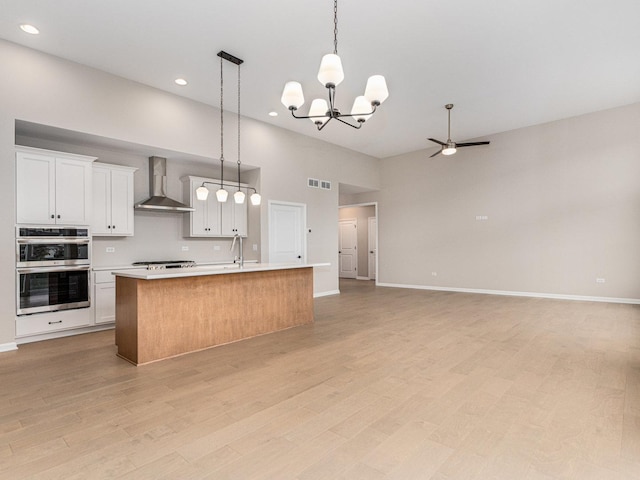 kitchen featuring stainless steel double oven, white cabinets, wall chimney exhaust hood, hanging light fixtures, and a center island with sink