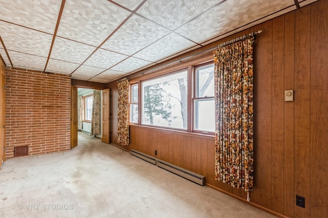 carpeted empty room with wood walls, a drop ceiling, brick wall, and a baseboard heating unit