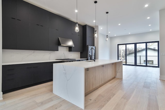 kitchen with sink, wall chimney range hood, backsplash, pendant lighting, and a center island with sink