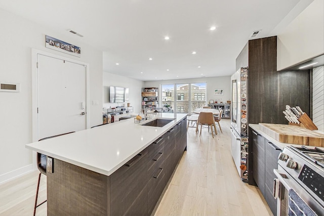 kitchen featuring a large island with sink, sink, stainless steel stove, light wood-type flooring, and dark brown cabinets