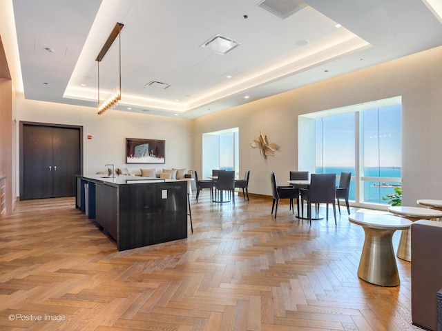 kitchen with light parquet flooring, decorative light fixtures, a tray ceiling, a center island with sink, and a water view