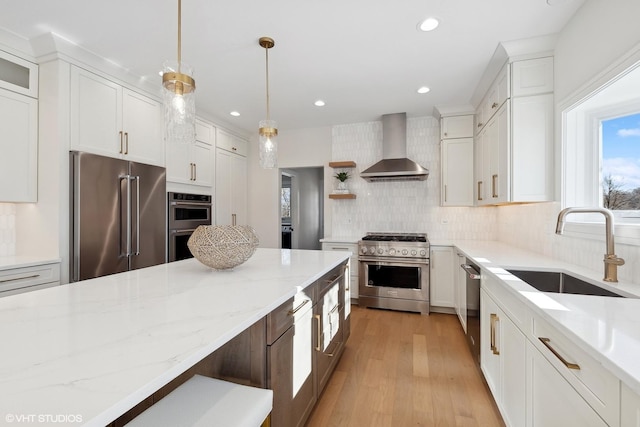 kitchen featuring wall chimney range hood, sink, high end appliances, white cabinets, and decorative light fixtures