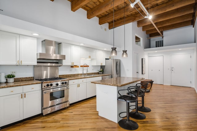 kitchen with beamed ceiling, a center island, appliances with stainless steel finishes, wall chimney range hood, and white cabinets