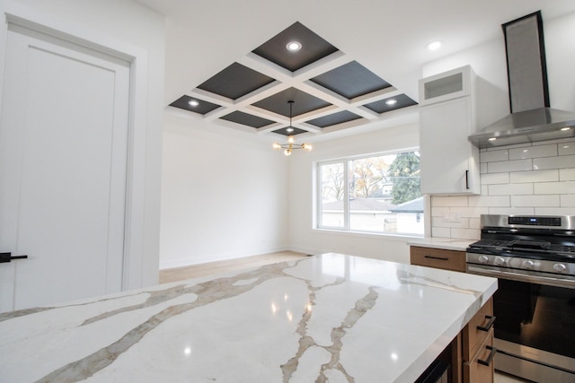 kitchen featuring light stone counters, wall chimney exhaust hood, coffered ceiling, stainless steel gas stove, and white cabinetry