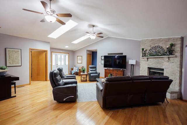 living area featuring light wood finished floors, a brick fireplace, ceiling fan, vaulted ceiling with skylight, and baseboards