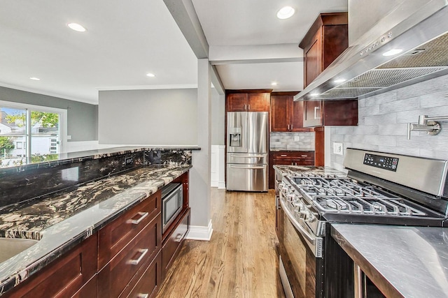 kitchen featuring wall chimney exhaust hood, dark stone countertops, appliances with stainless steel finishes, tasteful backsplash, and light hardwood / wood-style floors