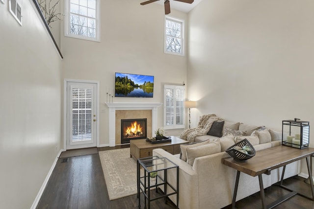 living room with a fireplace, a towering ceiling, ceiling fan, and dark wood-type flooring